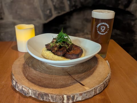 Seared pork belly over creamy grits with crispy herbs, beside a pint of Concordance Ferments beer