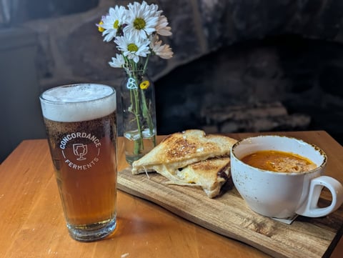 Grilled cheese on a board with a cup of tomato soup and a pint of beer by the stone hearth